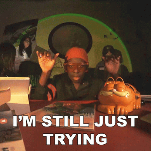 A Man Sitting At A Table With A Garfield Figurine And The Words I 'M Still Just Trying GIF