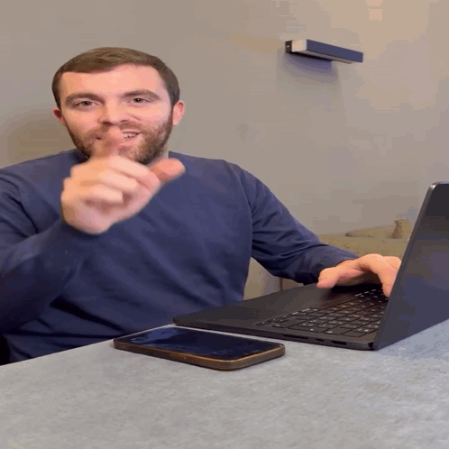 A Man Sitting At A Table With A Laptop And Cell Phone GIF