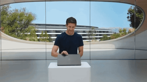 A Man Sitting At A Table With A Laptop In Front Of A Large Window GIF