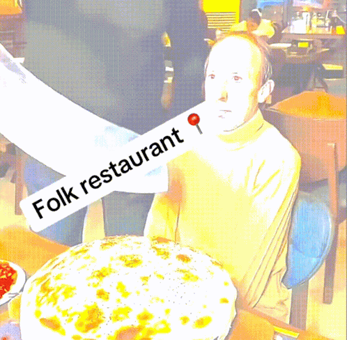 A Man Sitting At A Table With A Large Pizza And A Sign That Says Folk Restaurant GIF