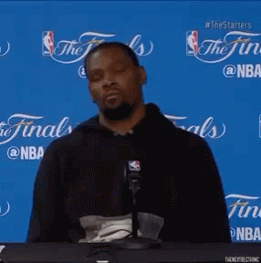 A Man Sitting At A Table With A Microphone In Front Of A Blue Background That Says The Finals On It GIF