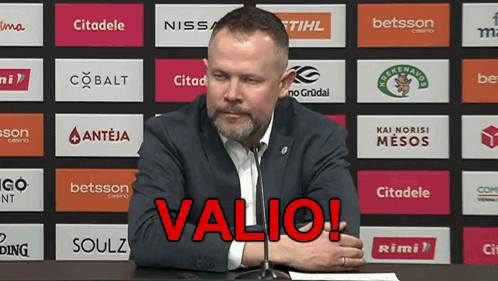 A Man Sitting At A Table With A Microphone In Front Of A Wall With Logos For Various Companies Including Valio GIF
