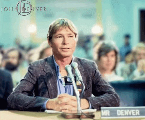 A Man Sitting At A Table With A Sign That Says Mr Denver On It GIF