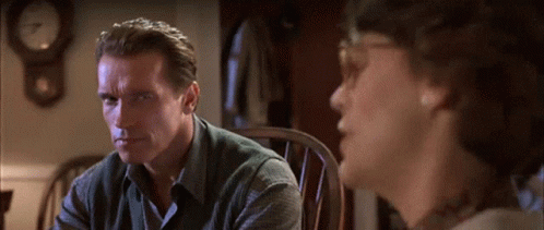 A Man Sitting In A Chair Talking To Another Man With A Clock In The Background GIF