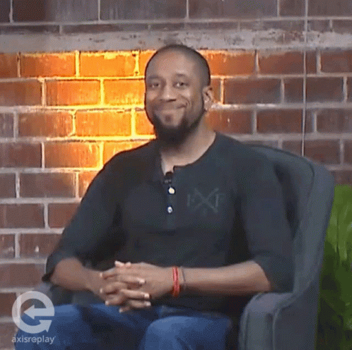A Man Sitting In A Chair With A Brick Wall Behind Him And A Logo For Axisreplay On The Bottom GIF