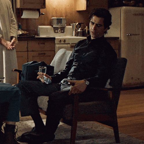 A Man Sitting In A Chair With A Glass Of Water In His Hand GIF