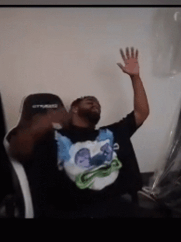 A Man Sitting In A Chair With His Hand In The Air Wearing A Shirt That Says ' Crysis ' On It GIF