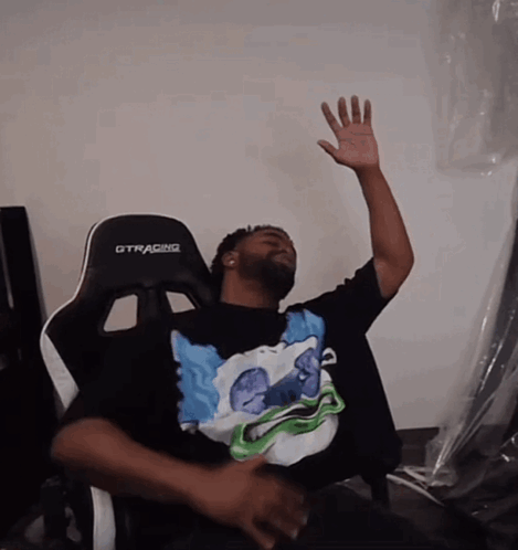 A Man Sitting In A Gtracing Chair With His Hand Up In The Air GIF