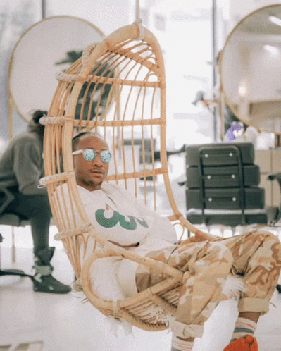 A Man Sitting In A Wicker Chair Wearing Sunglasses And A White Shirt With A Green Leaf On It GIF