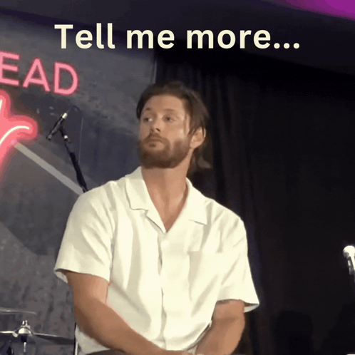 A Man Sitting In Front Of A Neon Sign That Says " Tell Me More " GIF