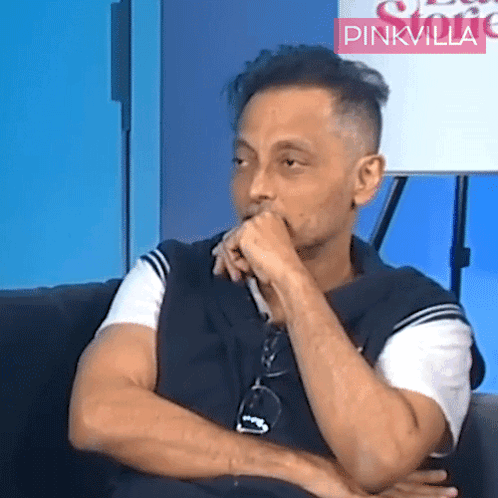 A Man Sitting In Front Of A Sign That Says Pinkvilla On It GIF