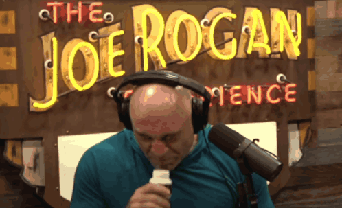 A Man Sitting In Front Of A Sign That Says The Joe Rogan Fence GIF