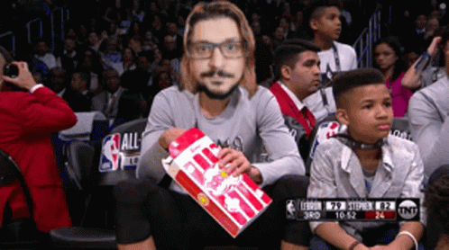 A Man Sitting In The Stands With A Mask On His Face Holding A Bag Of Popcorn GIF