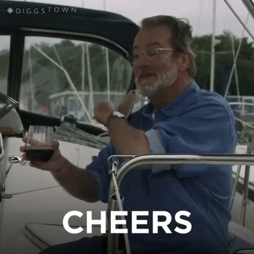 A Man Sitting On A Boat Holding A Glass Of Wine With The Word Cheers Behind Him GIF