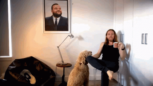 A Man Sitting On A Chair Next To A Dog Holding A Coffee Mug GIF
