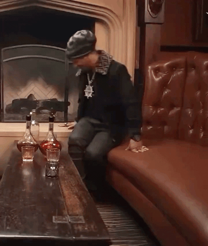 A Man Sitting On A Couch In Front Of A Fireplace With Two Bottles Of Liquor On The Table GIF