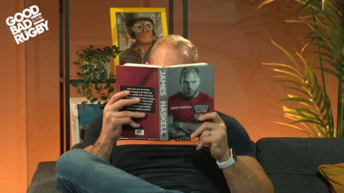 A Man Sitting On A Couch Reading A Book Called James Haskell GIF