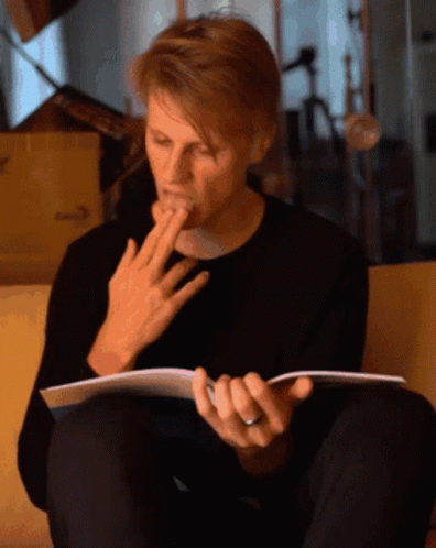 A Man Sitting On A Couch Reading A Book With His Hand On His Mouth GIF