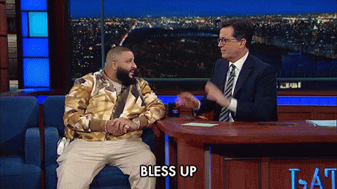 A Man Sitting On A Couch Talking To A Man In A Suit And Tie With The Words Bless Up On The Table GIF
