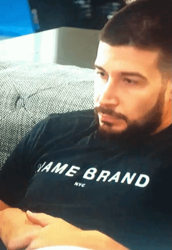 A Man Sitting On A Couch Wearing A Black Shirt That Says Name Brand Nyc GIF