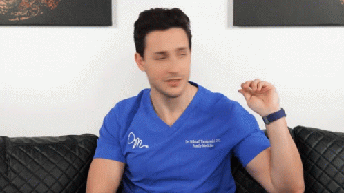 A Man Sitting On A Couch Wearing A Blue Scrub Top That Says Dr. Michał Tuchinski GIF