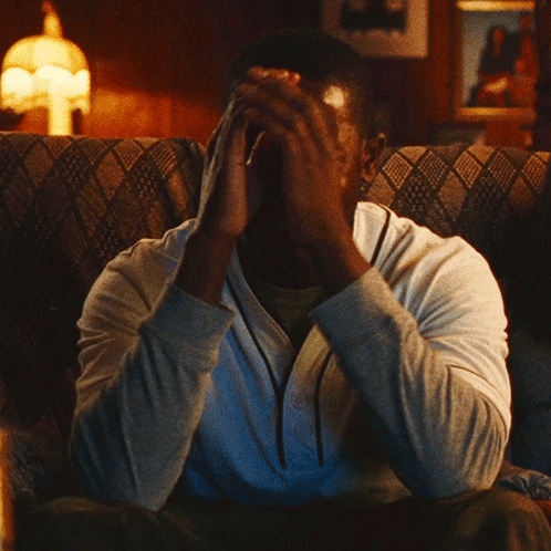 A Man Sitting On A Couch With His Hand On His Face GIF