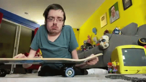 A Man Sitting On The Floor Holding A Skateboard With A Yellow Tv Behind Him GIF
