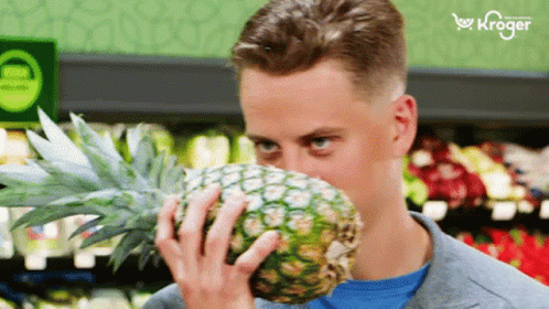 A Man Smells A Pineapple In A Grocery Store With A Kroger Logo In The Background GIF