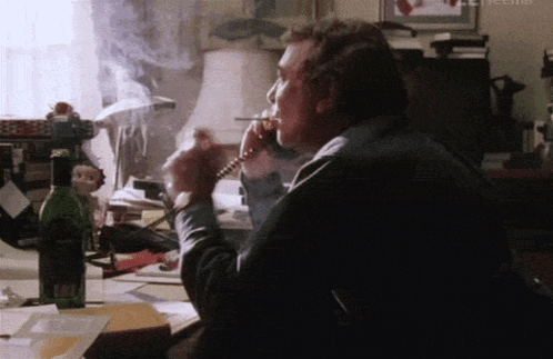 A Man Smoking A Cigarette While Talking On A Phone With A Bottle Of Wine In The Background GIF