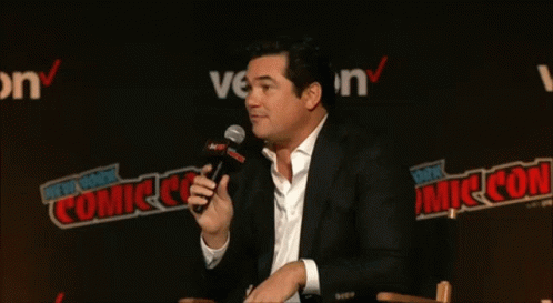 A Man Speaking Into A Microphone In Front Of A Wall That Says New York Comic Con GIF