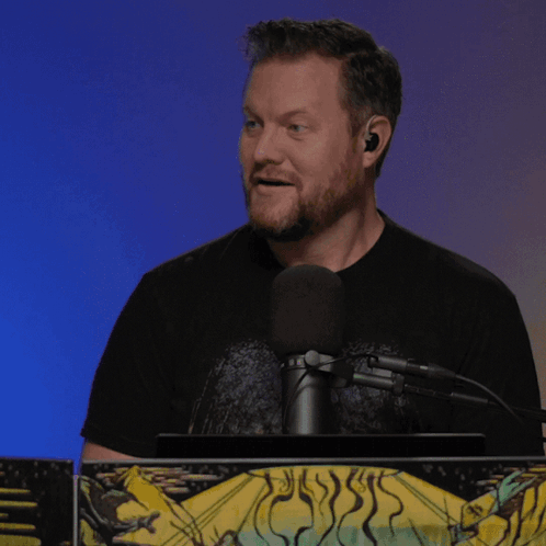 A Man Speaking Into A Microphone While Wearing Earbuds And A Black Shirt GIF