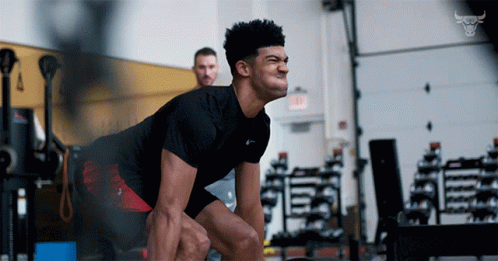 A Man Squatting In A Gym With A Bull Logo On The Wall Behind Him GIF