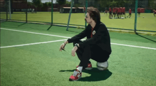 A Man Squatting On A Soccer Ball On A Field With A Team In The Background GIF