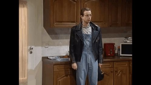A Man Standing In A Kitchen Wearing Overalls And A Leather Jacket GIF