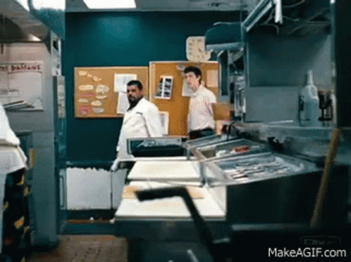A Man Standing In A Kitchen With A Sign On The Wall That Says Delicious GIF