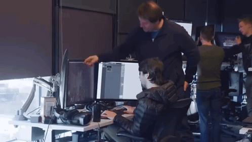 A Man Standing Next To A Man Sitting At A Desk Looking At A Computer Screen GIF