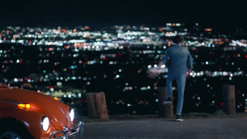 A Man Standing Next To An Orange Car Looking At A City GIF