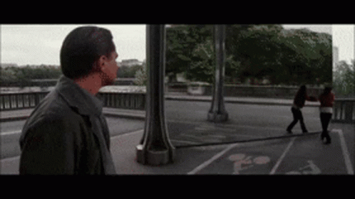 A Man Standing On A Bridge Looking At A Woman Walking On The Sidewalk GIF