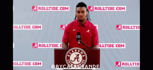 a man stands at a podium in front of a microphone wearing a red shirt with the letter a on it