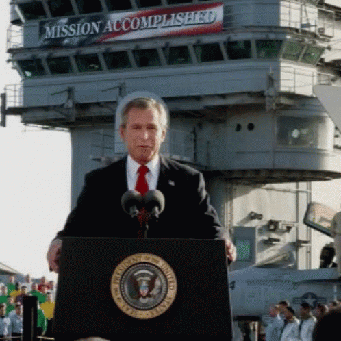 A Man Stands At A Podium In Front Of A Sign That Says Mission Accomplished On It GIF