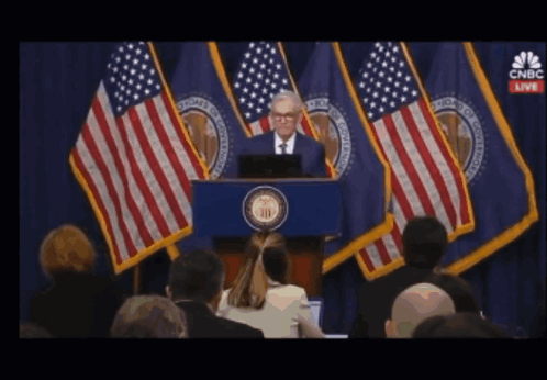 A Man Stands At A Podium In Front Of American Flags With Cnbc Live On The Screen Behind Him GIF