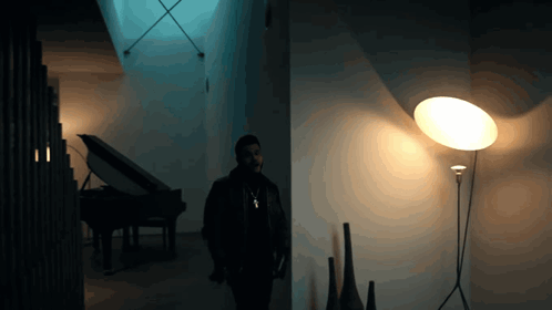A Man Stands In A Dark Room With A Lamp And A Piano GIF