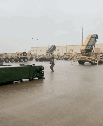 A Man Stands In A Parking Lot With Military Vehicles And A Container That Says ' Army ' On It GIF