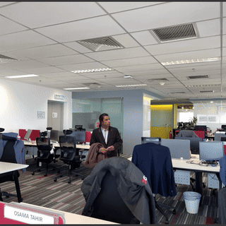 A Man Stands In An Office With A Sign That Says Osama Tahir On It GIF
