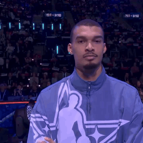 A Man Stands In Front Of A Crowd Wearing A Blue Jacket With A Basketball Player On It GIF