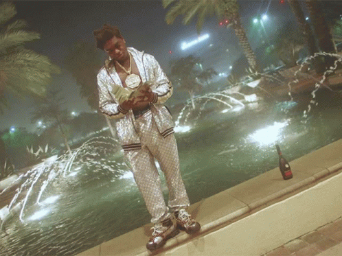 A Man Stands In Front Of A Fountain With A Bottle Of Champagne In Front Of Him GIF