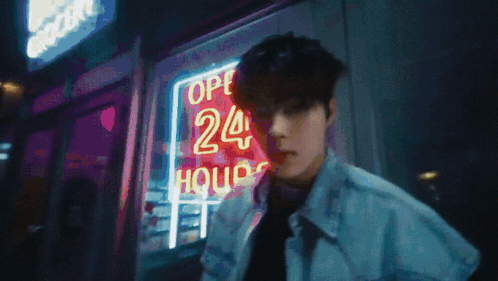 A Man Stands In Front Of A Neon Sign That Says 24 Hours GIF