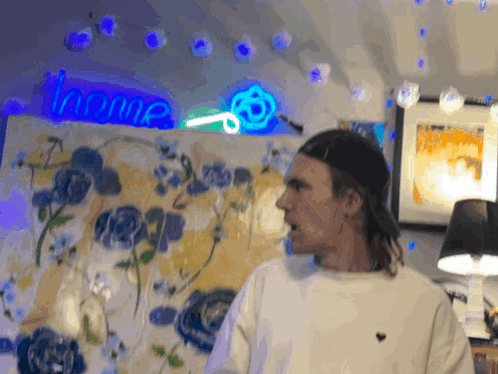 A Man Stands In Front Of A Painting With A Neon Sign That Says Home Behind Him GIF
