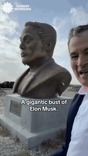 A Man Stands In Front Of A Statue Of Elon Musk And Says A Gigantic Bust Of Elon Musk GIF