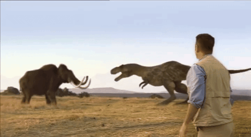 A Man Stands In Front Of A T-Rex And A Mammoth In A Field GIF